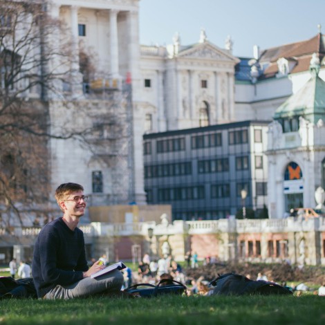 A student sits on the grass of an urban park on a sunny day and looks up from a book, smiling.