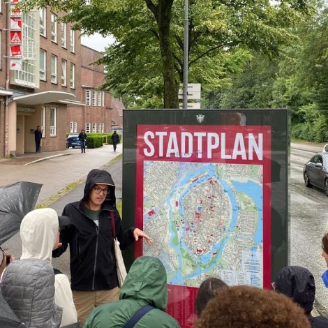 The group gathers outdoors around the professor, who is speaking while standing next to a map. Many of the participants are holding umbrellas. Behind them are a large brick building, trees, and a road with cars passing by.
